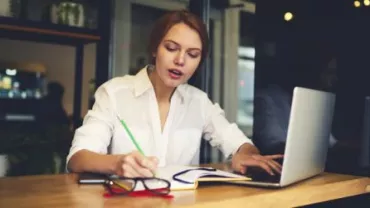 Woman writing notes and using a laptop at a cafe.
