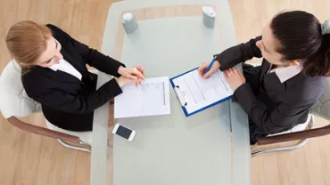Two businesswomen at a glass table reviewing documents.