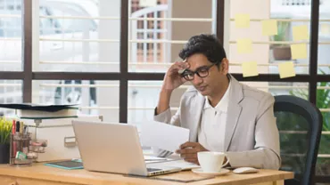 Man at desk reading paper by laptop with post-it notes on the window.