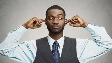 Man in vest and tie closes ears with fingers, looking to the side.