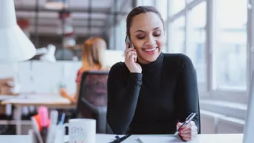 Smiling woman on phone at desk, taking notes in a bright office.