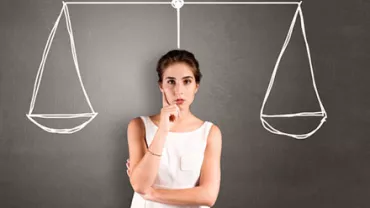 Woman pondering, standing in front of sketched balance scales on a blackboard.
