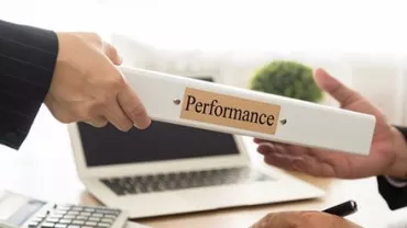 Two people exchanging a binder labeled 'Performance' at a desk with a laptop and calculator.