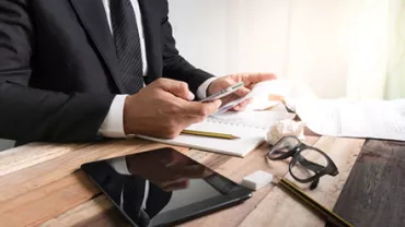 Businessman in suit using smartphone at a desk with tablet and glasses.