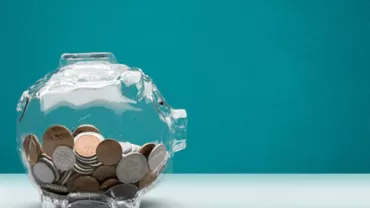 Clear piggy bank filled with various coins against a teal background.