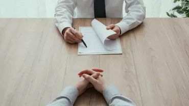 Two people sitting at a table, one holding a pen and paper, in an office setting.