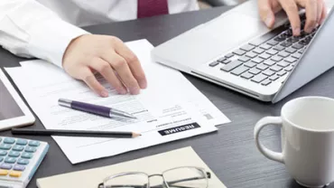 Man in office working on documents and laptop with a calculator, mug, and glasses on the desk.
