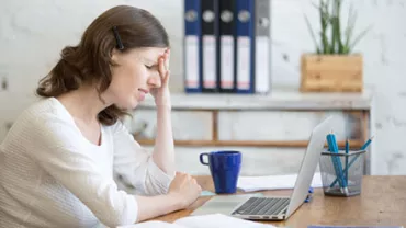Woman holding head in frustration at desk with laptop and coffee.