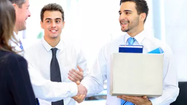 A man holding a box is smiling and shaking hands with another man in an office setting.