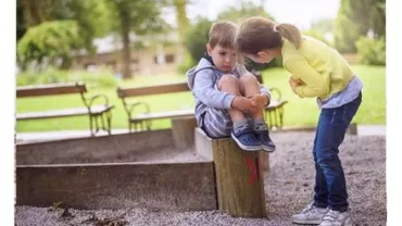 A boy sitting on a wooden post looking sad as a girl leans over to talk to him in a park setting.