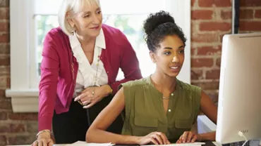 Older woman helping a young woman at a computer in an office.