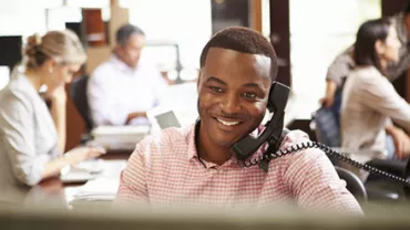 Man smiling while talking on the phone in a busy office.