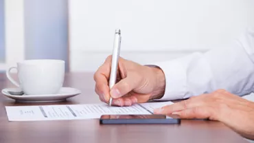 Person signing a paper at a desk with a pen and smartphone nearby.