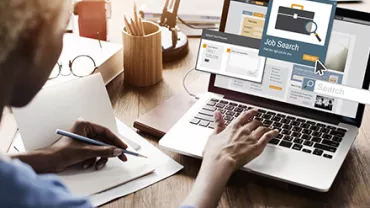 Person writing notes while using a laptop for job searching.
