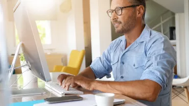 Man in glasses working at a desktop computer in a bright room.