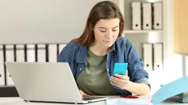 Woman in denim shirt checks phone at desk with laptop.