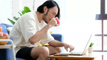 Man in a white shirt drinking coffee while working on a laptop.