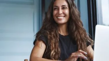 A woman with long hair smiles while working on a laptop by a window.