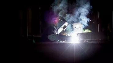 Welder at work with bright sparks and smoke in a dimly lit workshop.