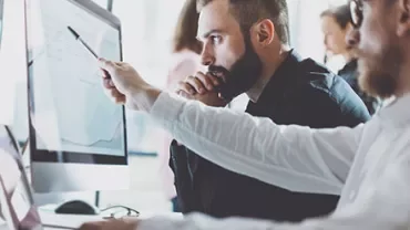 Two men working on desktop computers, discussing data on a screen in a modern office.
