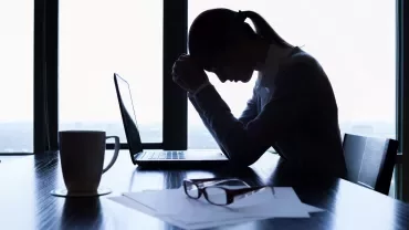 Silhouette of a person at a desk, head bowed near a laptop with a mug and papers.