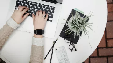 Hands typing on a laptop beside a plant and glasses on a white table.