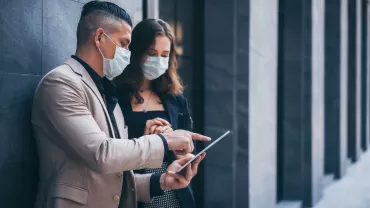 Two people in masks discussing on a tablet outdoors.