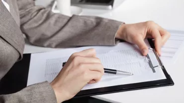 Person in suit reviewing documents on clipboard at desk.