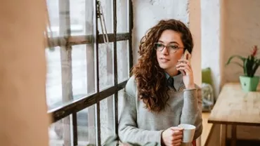 Woman with curly hair on phone, holding a mug, sitting by a window in a cozy cafe.