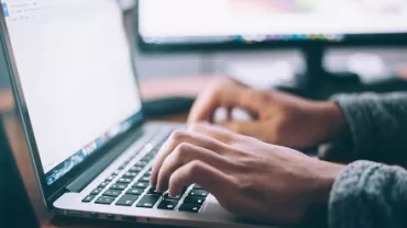 Person typing on a laptop with a second monitor in the background.