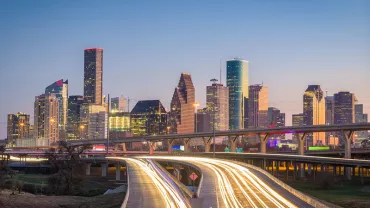 City skyline at dusk with illuminated skyscrapers and light trails on the highway.