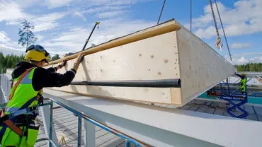 Construction worker guides a large wooden panel into place at a building site under a clear sky.