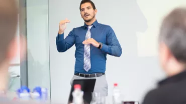 Man giving a presentation in a blue shirt and tie, gesturing with hands.