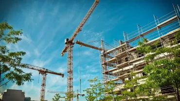 Three cranes towering over a building under construction with scaffolding against a blue sky.