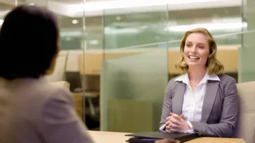 Two people in a business meeting, smiling and talking across a table.