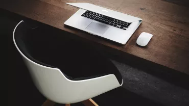 Open laptop and mouse on a wooden desk with a modern chair.