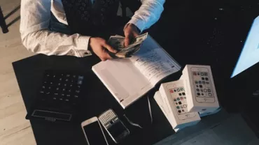 Person counting money at a desk with phones and a notebook.