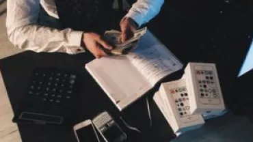 Person counting cash at a desk with electronic devices and a notebook.