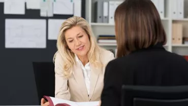A woman in a beige blazer discusses a document with another woman in an office.