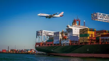 A cargo ship in port with colorful containers and an airplane flying overhead under a clear blue sky.