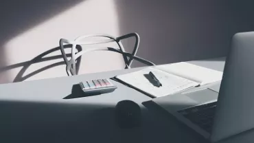 A laptop, notepad, pen, and calculator on a sunlit desk.