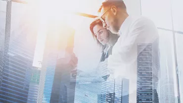 Double exposure of two business professionals working on a tablet with skyscrapers and sunlight in the background.