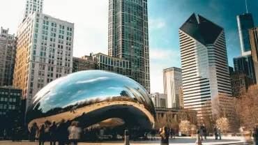 The Bean sculpture in a city plaza reflecting buildings.