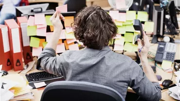 Person at desk surrounded by sticky notes and binders, holding a phone.