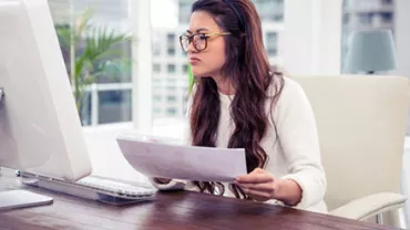 Woman in glasses working at a computer, holding documents.