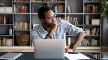 Man sitting at desk with laptop, thinking, bookshelf in background.