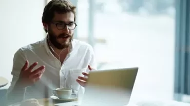 Man wearing headphones, engaged in a video call on a laptop, sitting at a cafe with a cup of coffee.