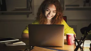 Woman with red hair smiling at a laptop screen in a dimly lit room.