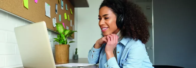 Woman with headphones smiling at laptop, working at home office desk.