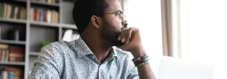 Man in patterned shirt looking thoughtfully out a window near a laptop.